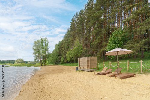 Pine and birch trees border a sandy beach with wooden sunbeds and an umbrella. A calm river flows near a forest under a cloudy summer sky.