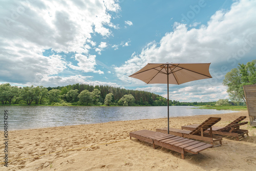 Wooden sunbeds and an umbrella stand on a sandy beach by a wide river. Pine and willow trees grow on the opposite green bank under a cloudy sky.