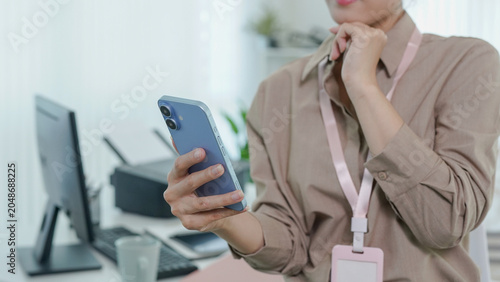 A professional woman in a brown shirt holds her smartphone while resting her chin on her hand at her office desk.