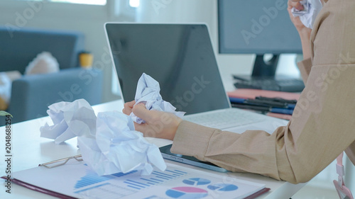 A close-up view depicts a woman's hand holding crumpled paper over a desk with financial charts and a laptop.