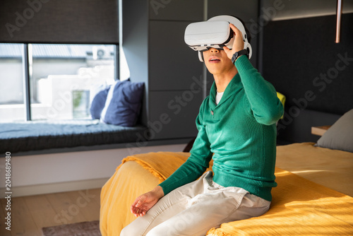 White VR headset resting on bed edge with yellow blanket, gray pillow, bright window seat