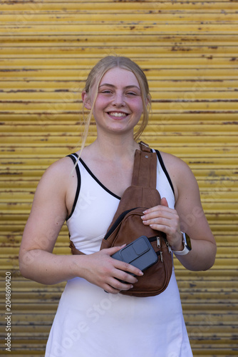 Woman in 20s posing by yellow shutter wearing white dress holding phone brown sling bag smartwatch