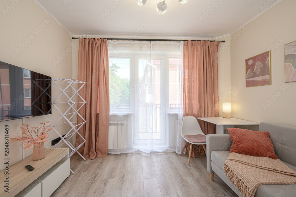 Fototapeta premium cozy living room with a grey sofa, peach curtains, a white desk, and a TV unit; light wood flooring, abstract art, and a folding drying rack add warmth and functionality