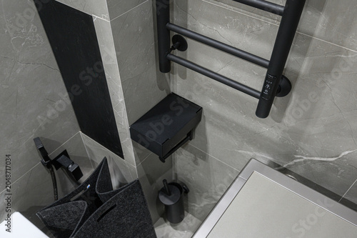 Modern bathroom corner with grey marble tiles, matte black heated towel rail, floating shelf, toilet brush holder, and felt laundry baskets in monochrome design