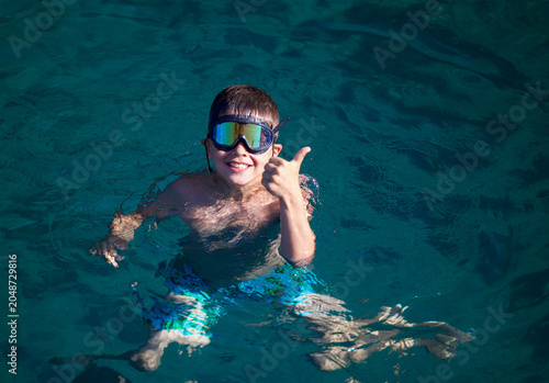 A young person wearing mirrored swim goggles treads water in deep turquoise sea, smiling broadly and giving a thumbs-up gesture. Sunlight reflects across the