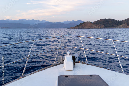 View from the bow of a white motorboat looking out over rippling blue water toward a distant mountainous coastline under a soft pastel dusk sky. Stainless