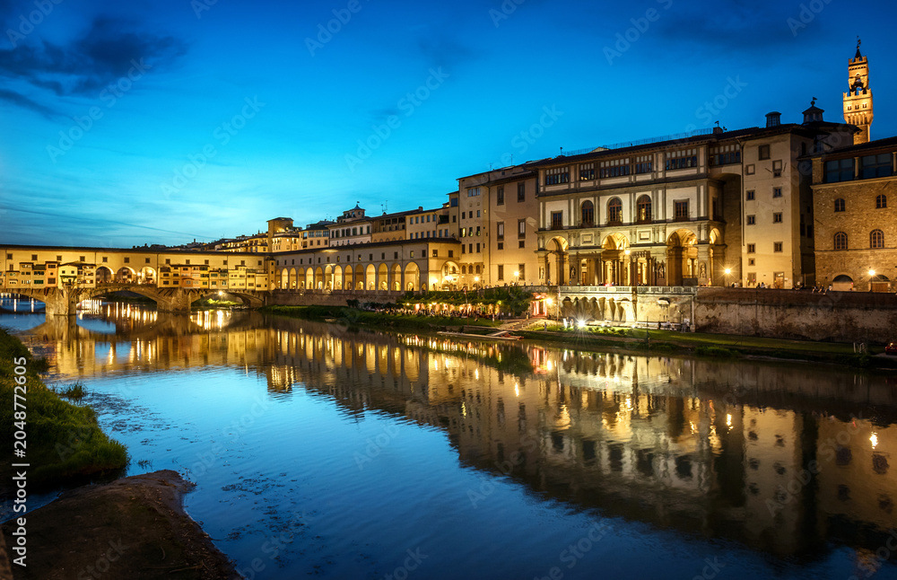 Fototapeta premium Night view of Florence, Italy, featuring the illuminated Ponte Vecchio bridge and the famous Uffizi Gallery along the Arno River. Historic Renaissance architecture reflected in calm water 