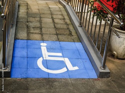 A view of an inclusive urban infrastructure design, featuring a ramped pedestrian entrance flanked by steel guardrails and disability-friendly signage under daylight.