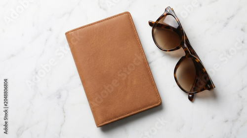 Blank brown leather passport holder or notebook mockup with tortoiseshell sunglasses on white marble background, top view