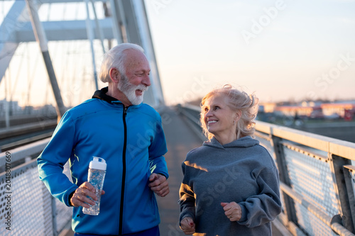 Happy senior couple jogging together on a modern bridge at sunrise, smiling and enjoying an active healthy lifestyle. Fitness, wellness, longevity and positive aging concept