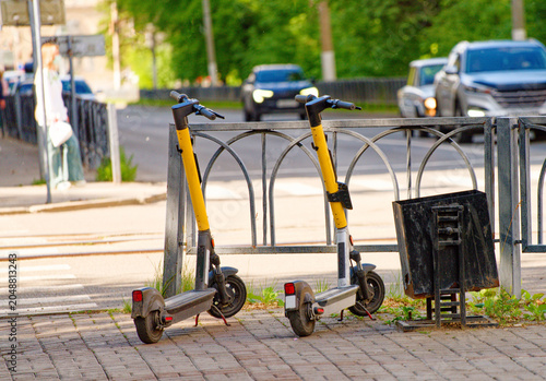 Electric scooters standing by a metal fence on a sunny urban street. Modern public transportation, mobility and city life concept with moving traffic in the background. Photo