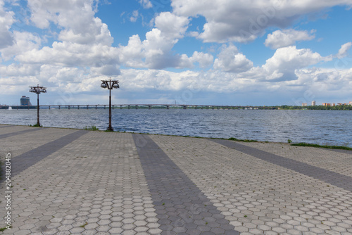 Wide paved riverside promenade with calm water, cloudy sky and city waterfront view