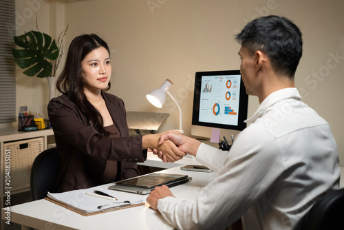 Asian Business Professionals Shaking Hands Across Desk in Office with Data Charts on Monitor