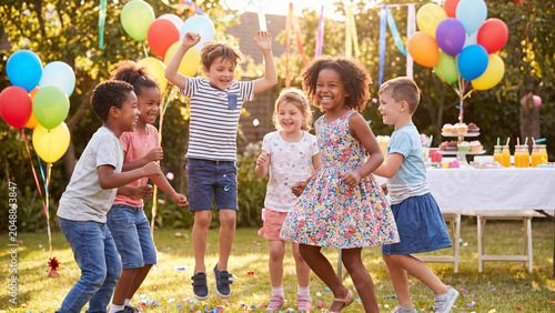 Group of diverse children jump with excitement during an outdoor birthday celebration in a garden, surrounded by floating colorful balloons, festive party decorations, and a decorated table.
