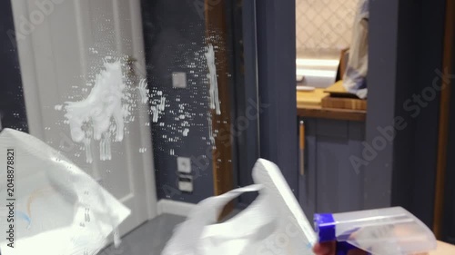 A teenage girl sprays cleaner and wipes a mirrored wardrobe door with a paper towel during household chores in a home hallway.