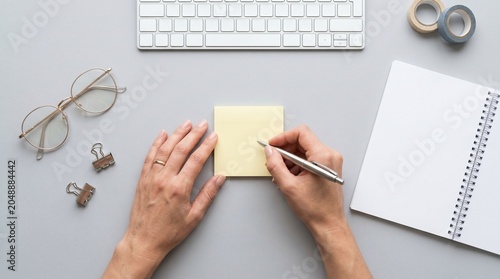 Person writing note on sticky pad at minimalist desk with glasses and coins