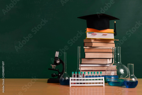Education concept from school to college and university, microscope, textbooks, chemistry glassware and mortarboard on table against chalkboard. Blank space, copy space.
