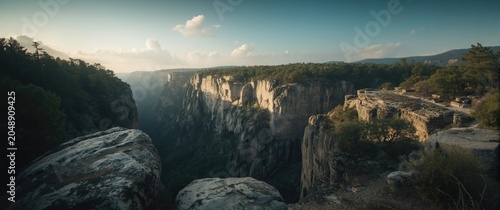 High angle view of a deep gorge lined with imposing cliffs and large foreground rocks beneath a lightly clouded blue sky copy space
