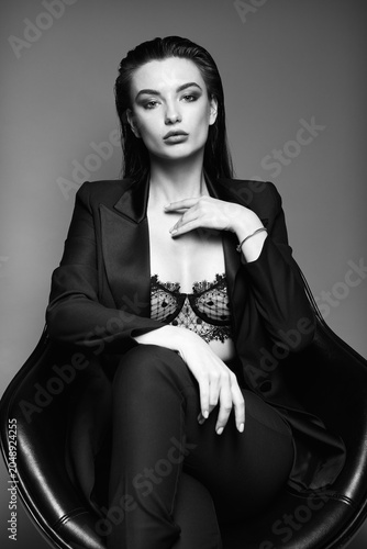 Studio fashion: gorgeous young woman dressed in black suit (jacket and pants) and bra. Portrait of pretty girl sitting on chair against gray background. Black and white