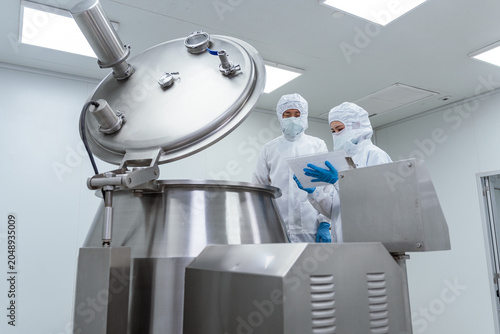 Two scientists in protective gear inspect mixing equipment in. Professional scientists in cleanroom suits inspecting pharmaceutical manufacturing processes and operating industrial mixing equipment.