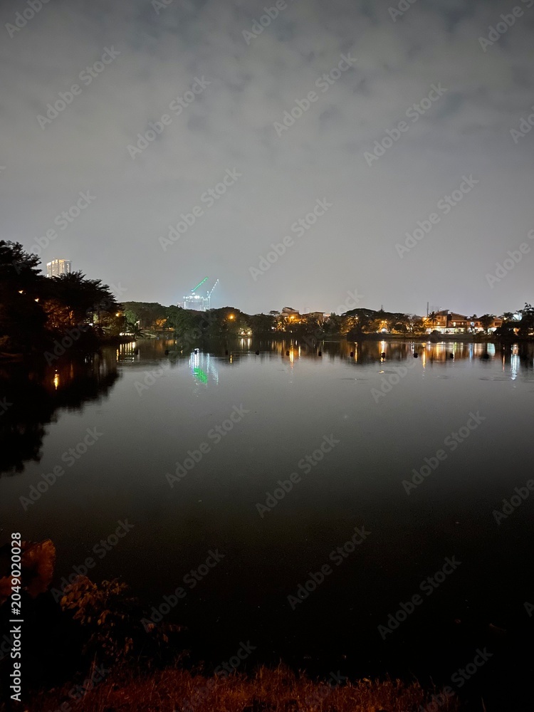 Fototapeta premium Night landscape of a calm lake reflecting a house and white clouds on the water surface. Mirror-like reflection creates a peaceful scenic image with symmetry, tranquility, and natural beauty.