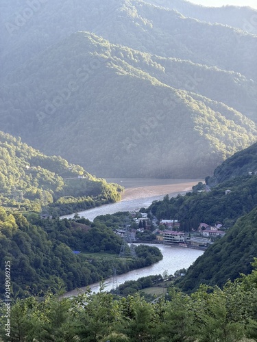 A scenic view of a winding river surrounded by lush green mountains. The landscape features a small village nestled in the valley, with soft sunlight illuminating the scene.