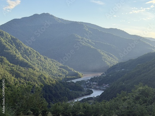 A scenic view of a winding river surrounded by lush green mountains under a clear blue sky. The landscape showcases natural beauty and tranquility.