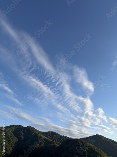 A clear blue sky with wispy clouds above green mountains. The landscape showcases natural beauty and tranquility in a serene outdoor setting.
