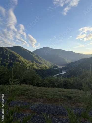A scenic view of mountains and a river under a blue sky with clouds. Lush greenery surrounds the landscape, creating a tranquil natural setting.
