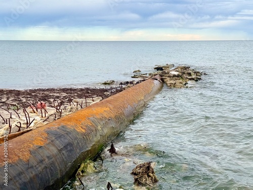 Fotografie An old rusted metal pipeline extending from the sandy shore into the choppy wate