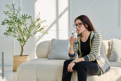 Obraz na płótnie Mature, serious woman with glass of water sitting on sofa, profile view