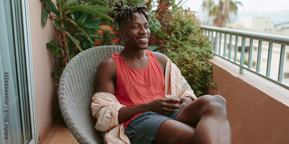 Fototapeta premium Smiling young man relaxing on a balcony chair in casual summer clothing for lifestyle banner or outdoor leisure background design