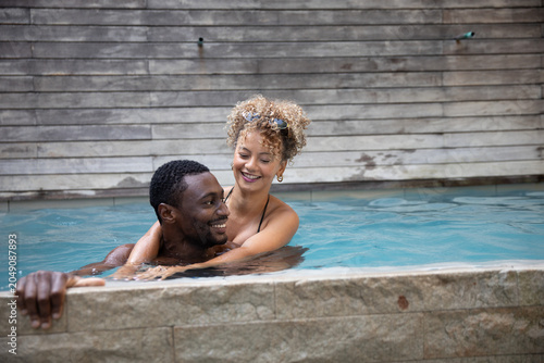 Diverse couple wearing bikini hugging while swimming at outdoor pool near stone coping, wooden wall