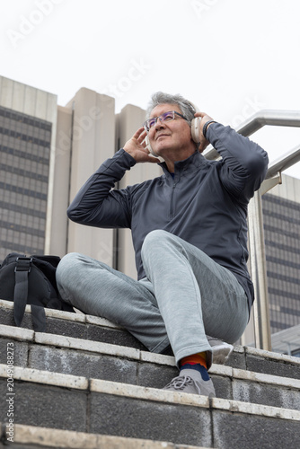 Billede på lærred Senior man sitting on stone steps in city plaza adjusting headset in athletic we