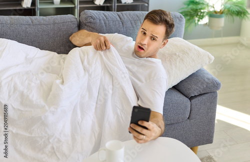 Fotografie Sleepy man checks alarm on phone on sofa