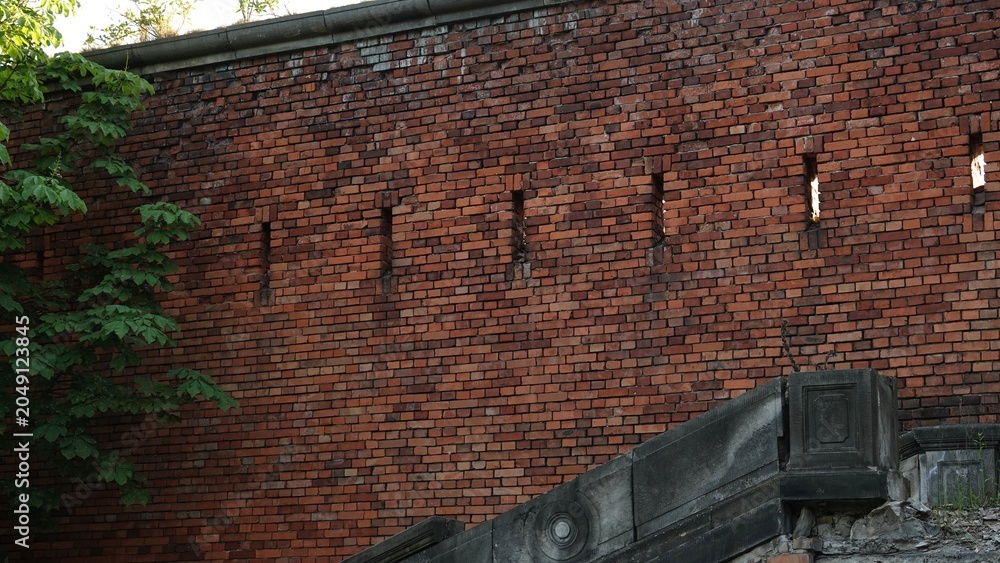 Fototapeta premium Monumentalny mur obronny z ciemnoczerwonej cegły z widocznymi otworami strzelniczymi, stanowiący fragment zabytkowej fortecy obronnej w otoczeniu zielonych liści drzew w letni dzień