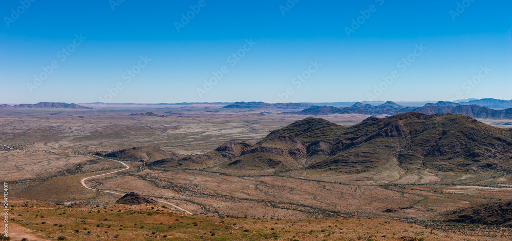 Obraz premium Aerial view of a semi arid Namibia landscape with low dark hills and mesas, a winding dirt road, and vast gravel plain under clear daylight, evoking solitude and scale.