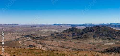 Aerial view of a semi arid Namibia landscape with low dark hills and mesas, a winding dirt road, and vast gravel plain under clear daylight, evoking solitude and scale.