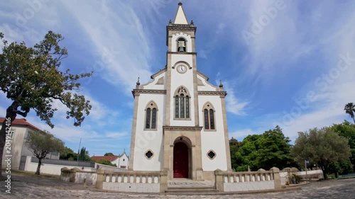 A symmetrical wide angle shot of the white facade of the historic parish church in Santa Maria de Lamas under a bright blue sky with wispy clouds. The bright daylight accentuates the textured stone de