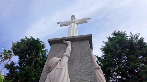 Low angle view of a stone monument featuring Jesus Christ with open arms atop a textured pedestal supported by sculpted figures. Bright daylight illuminates the scene against a blue sky.