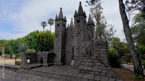 A detailed miniature castle built from small grey stone blocks stands in a green park under soft diffused daylight. The structure features corner towers battlements and a textured brick path.