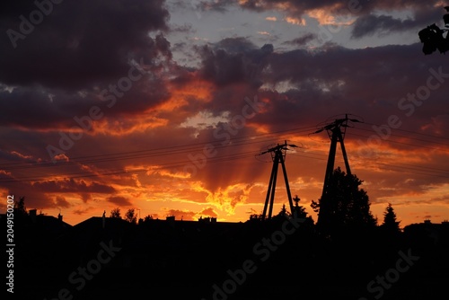 Power lines silhouette against dramatic sunset sky with dark clouds and fiery orange sunbeams over suburban houses