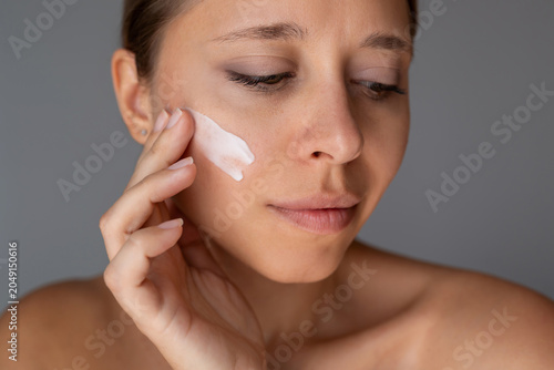 Close up of a young woman applying moisturizer cream on her cheek on gray background. Natural skin care routine for hydration, softness and healthy glowing complexion