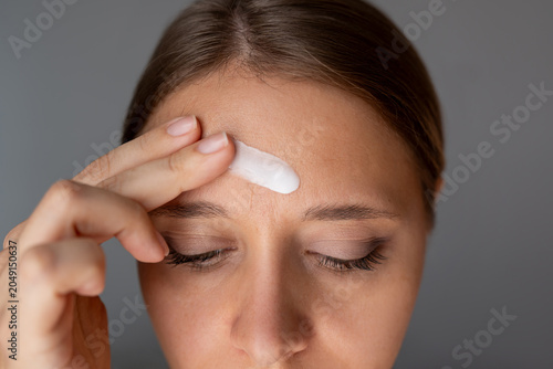 Young woman applying moisturizing cream on her forehead on gray background. Close up anti aging skin care routine for fine lines, wrinkles and facial hydration