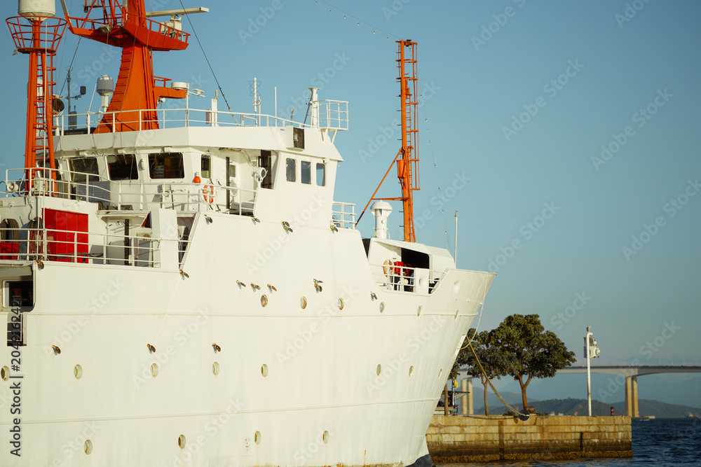 Naklejka premium Telephoto view of a white ship bow and bridge with orange masts moored beside a pier, water and a long bridge visible in warm sunlight