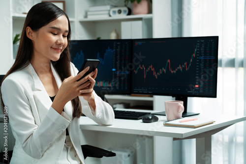 Quadro su tela Smiling young Asian businesswoman with happy face looking on smartphone in profitable of exchange stock market graph at modern office
