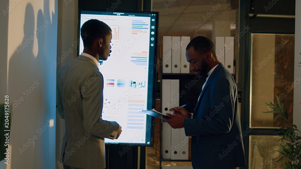 Fototapeta premium Manager debriefing employee in front of digital screen, showing him important research statistics before getting back to work. Businessmen in office during nightshift brainstorming business insights