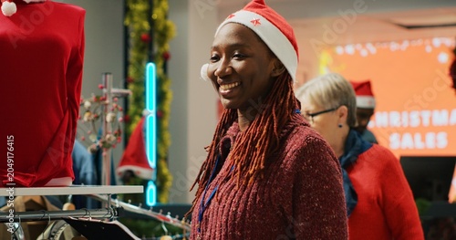 Fotografía Clothing store manager wearing festive Santa hat holding clipboard in mall shop inspecting rack of clothes and greeting customers