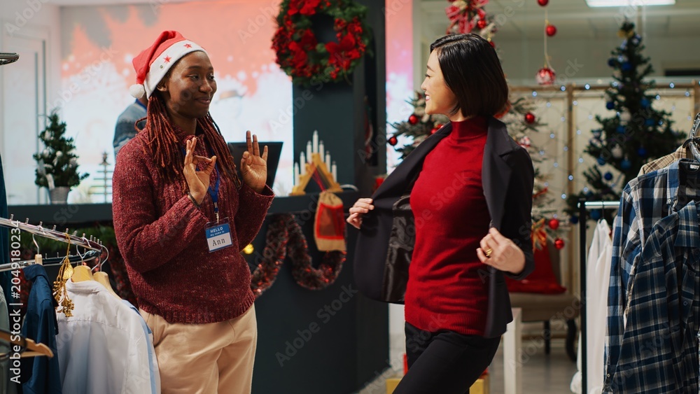 Obraz premium Asian businesswoman doing pirouette while trying stylish blazer, asking retail assistant for opinion before making purchase. Customer and employee in Christmas decorated clothing shop