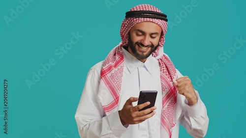Arab guy holding his mobile device, surfing across social media platforms and chatting online. Muslim with customary apparel uses smartphone to access global web, feeling excited on camera.